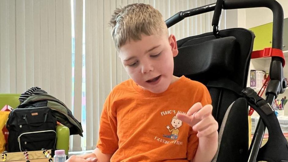 A boy with short blond hair in a pushchair wearing an orange t-shirt saying Isaac's Journey above a cartoon figure of a waving boy. The boy in the pushchair is looking downwards. Bags on a green sofa can be seen to his right, along with white vertical blinds. 