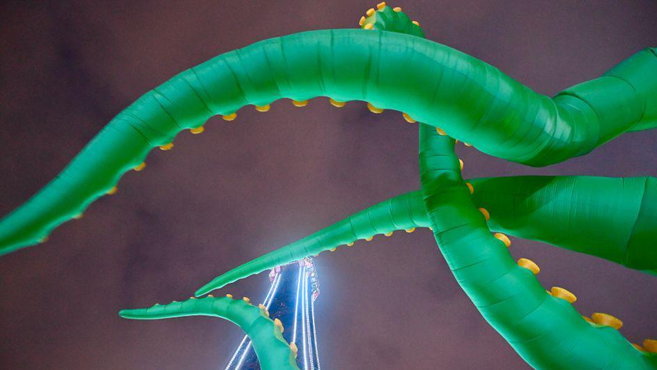 Four of the giant inflated tentacles viewed from underneath against the night sky. The tentacles are green with yellow nodules and Blackpool Tower, lit up in blue, can be seen through them towards the bottom of the picture