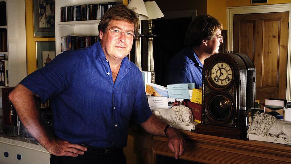 Edward Stourton is photographed at home in 2003. He is wearing a bright blue shirt and is resting his elbow on a fireplace