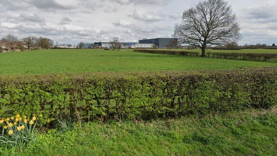 Pictured is a green field behind a hedgerow with some industrial-looking buildings at the back