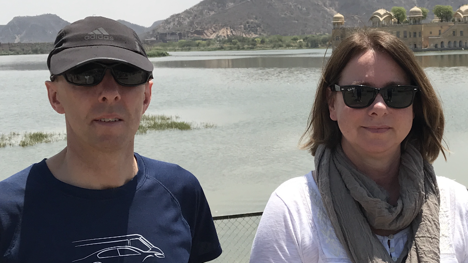 A man and woman stand in front of a body of water. The man - Chris - is in a blue Hendy T-shirt, a black cap and sunglasses. The woman - Carol - is wearing a white shirt, a khaki green scarf and sunglasses. She has chin-length brown hair. 