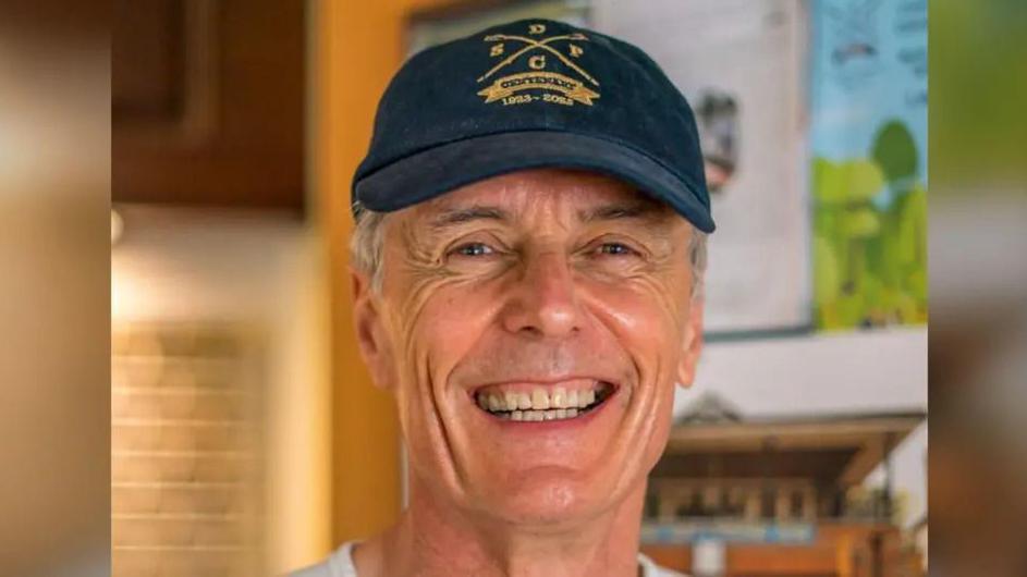 A man with white hair smiling at the camera. He is wearing a navy-coloured hat.