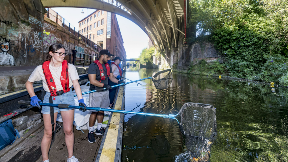 A woman with a large net picking litter up from a canal from a boat, with two other people next to her