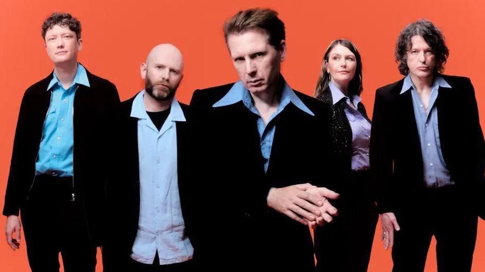 Indie rock band Franz Ferdinand in a publicity shot. The five members stand in a line all throwing different, fairly casual poses. All are wearing black suit jackets over coloured, wide collared button up shirts.