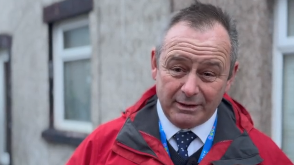 Dave Allen, PCC for Cumbria, on the dawn raid with the police. He is dressed in a suit with a red coat over the top of it. He has a blue lanyard around his neck.