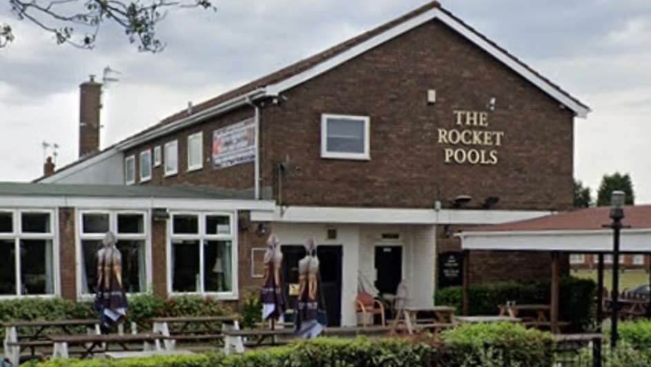 The pub has a garden at the front with tables and chairs outside and with its sign on a wall at the entrance to the pub.