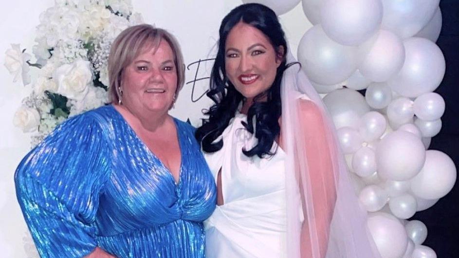 Susan McGowan with her niece Jade Campbell at her wedding - the two have their arms around each other and are standing in front of a white balloon archway. Ms McGowan has short blonde hair and is wearing an electric blue v-neck dress with long sleeves. Ms Campbell wears her long dark hair in waves with a white veil. Her wedding dress is brilliant white and has a keyhole neckline.