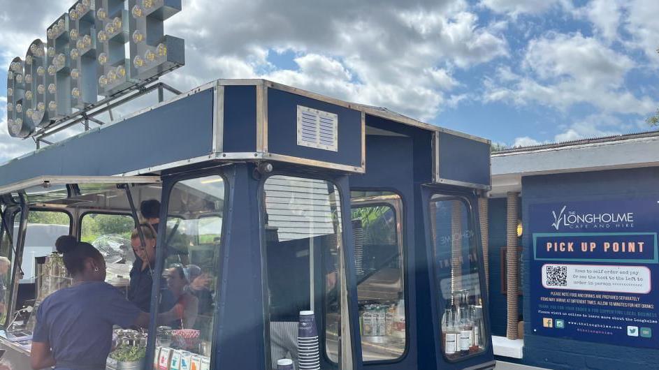 A blue mobile coffee van, by a sign, with one woman in front of it, and people working inside it.