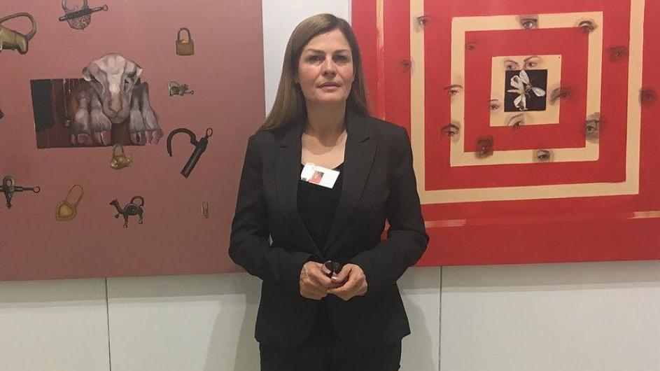Mehrangiz Imenpour is wearing a black suit with a vistor tag clipped to her shirt. She stands in front of a beige wall with two pieces of red art hanging on it.