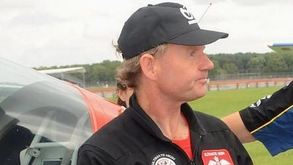 A man wearing a black pilot's suit and black cap, with a red shirt beneath. He is stood in front of a small aeroplane on an air field