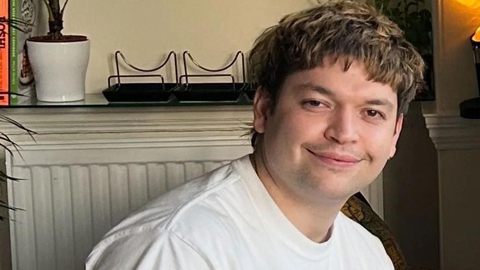 Luis Piovesana wearing a white t-shirt and sitting in a little room. He has brown eyes and medium length shaggy hair, which is dark brown with brighter highlights. He is clean shaven with a slight moustache. Behind him is a radiator with a shelf above it, displaying a potted plant and books.