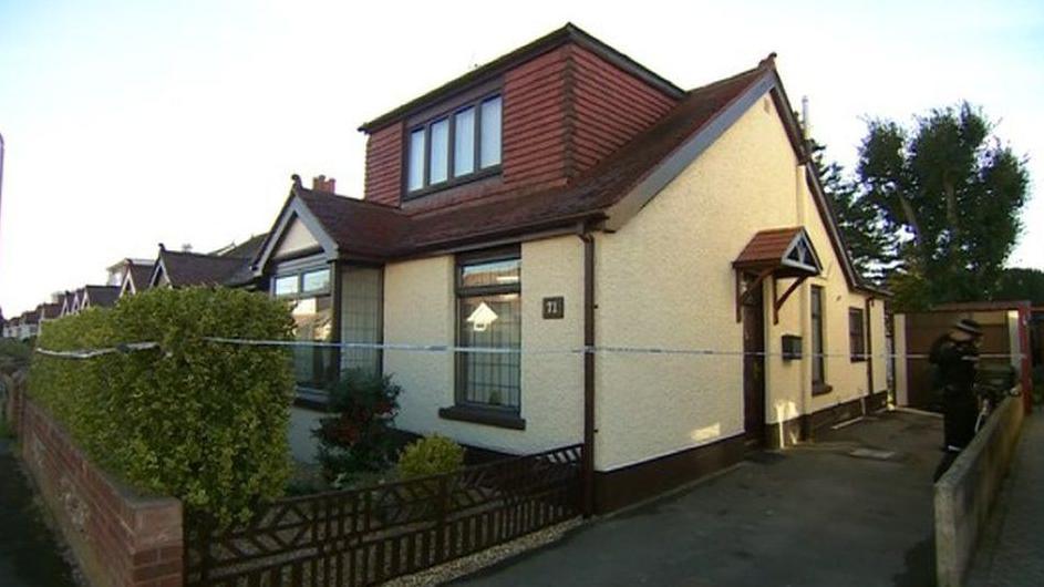 A two-storey house painted cream on the lower half with wooden panelling on the top half. The house is roped off by blue and white police tape.