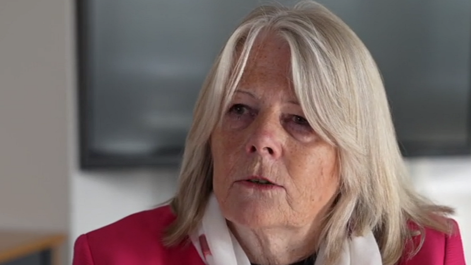 Penny Carpenter looks to the left of the camera, she is wearing a white scarf over a dark top and pink jacket. 