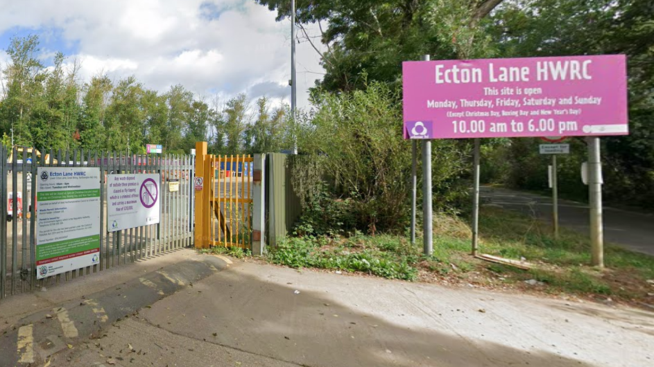 The outside of the waste centre - a big pink sign with "Ecton Lane HWRC" is by the entrance. There is a closed metal gate, which is the entrance of the site. There are trees in the background and blue skies above with some white cloud.