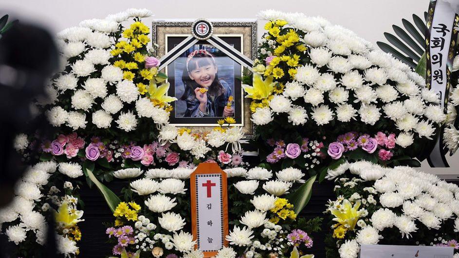 A framed portrait of Kim Ha-neul, smiling brightly with a pink hairband, mounted on a pile of white chrysanthemums.