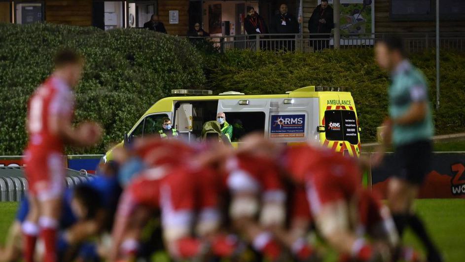 A yellow ambulance is seen in the background, behind a blurred picture of a rugby scrum