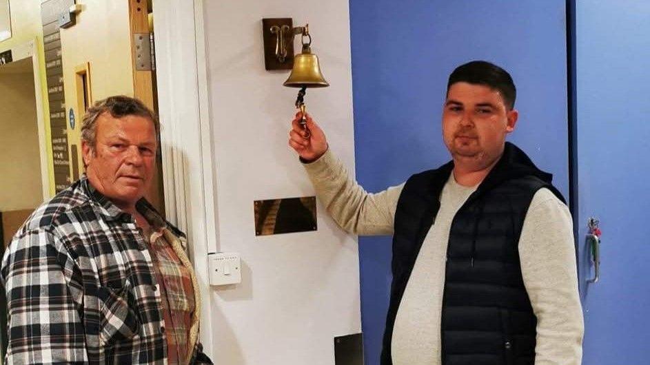 Liam Harvey wearing a grey sweatshirt underneath a black gilet. He is standing in hospital alongside his father, who is wearing a black and white plaid jacket. Liam is ringing a gold bell which marks the end of his treatment. 