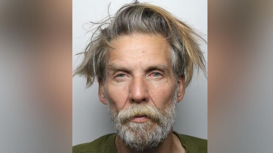 A police mugshot of a man in his late fifties. He has a narrow face, unkempt greying hair and a long beard. His eyes are blue and sullen.