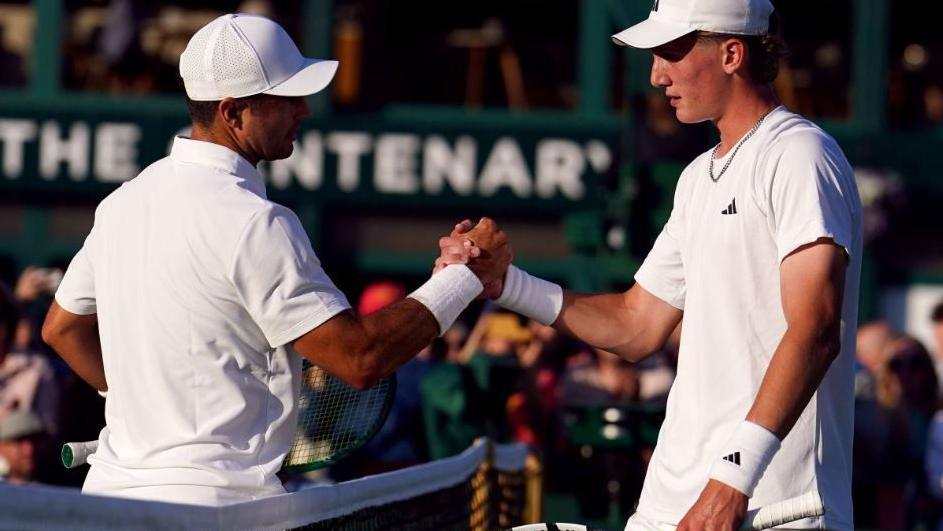 Two tennis players, both dressed all white, shake each others hands at the net on one of Wimbledon's grass courts. The crowd can be seen, blurred, in the background.