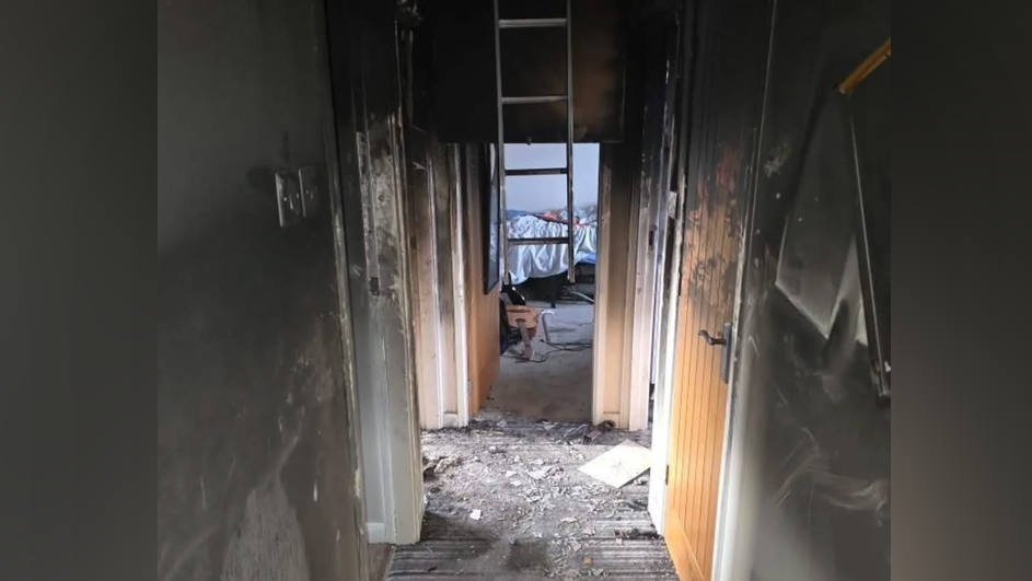 The hallway of the property with smoke blackened walls and debris on the floor. A ladder hangs halfway down from the entrance to the attic in the ceiling.