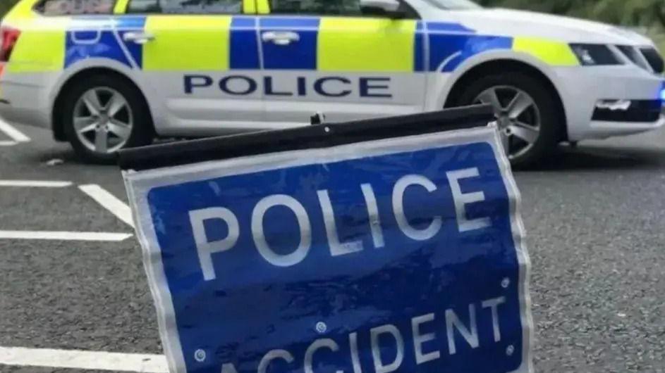 A police car parked on a road with a blue sign which reads 'police accident' positioned in the foreground.