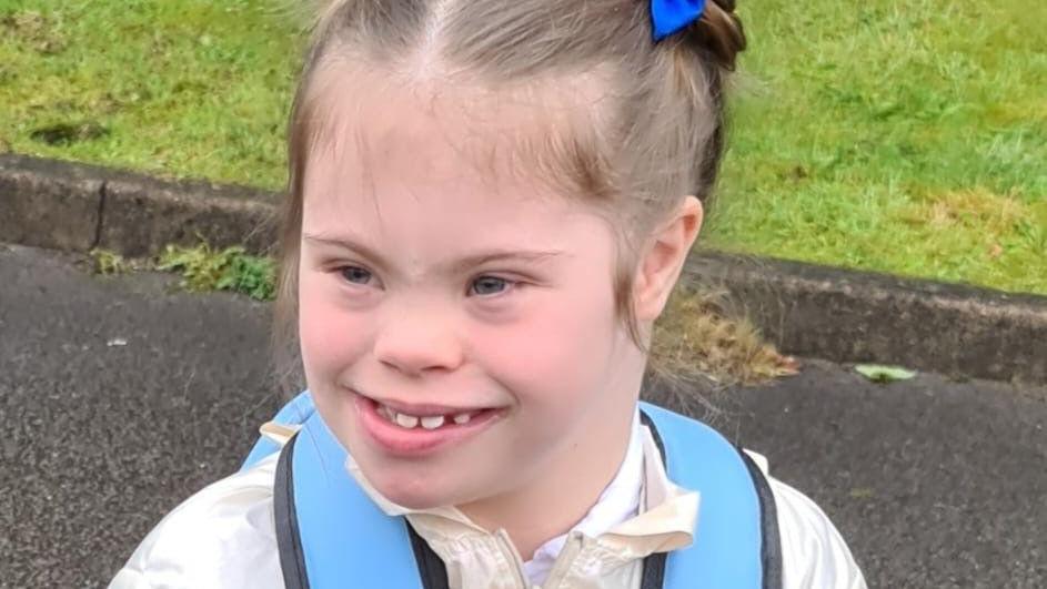 A young girl with brown hair, tied up in ponytails, smiles as she wears a cream jacket and blue rucksack