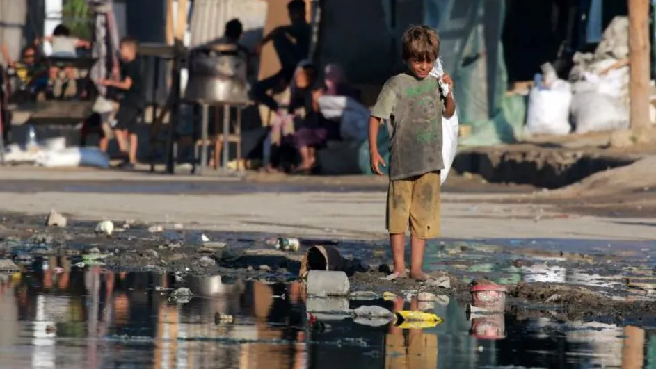 A child in dirty clothing stands in dirty water and rubbish with a plastic bag on his shoulder in Gaza