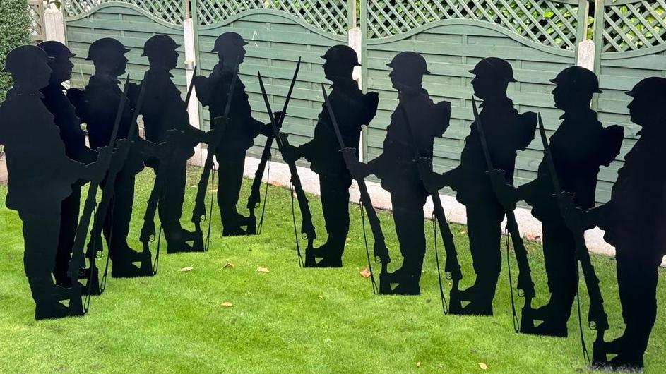 Ten black silhouette statues standing on a green lawn. The statues depict soldiers standing upright, each holding a rifle with the barrel pointing downward. Behind the statues is a green wooden fence.