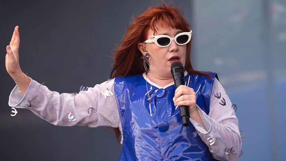 CMAT performing on stage wearing a blue dress and white sunglasses. She has red hair.