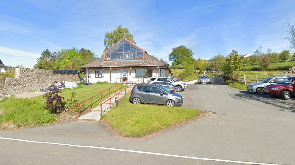 A white GP surgery with a brown tiled roof and glass window. There are concrete steps leading up to the front door, with red railings. Surrounding the step is green grass. To the right of the surgery is a car park with parked cars. In the background of the picture are green trees and a field.