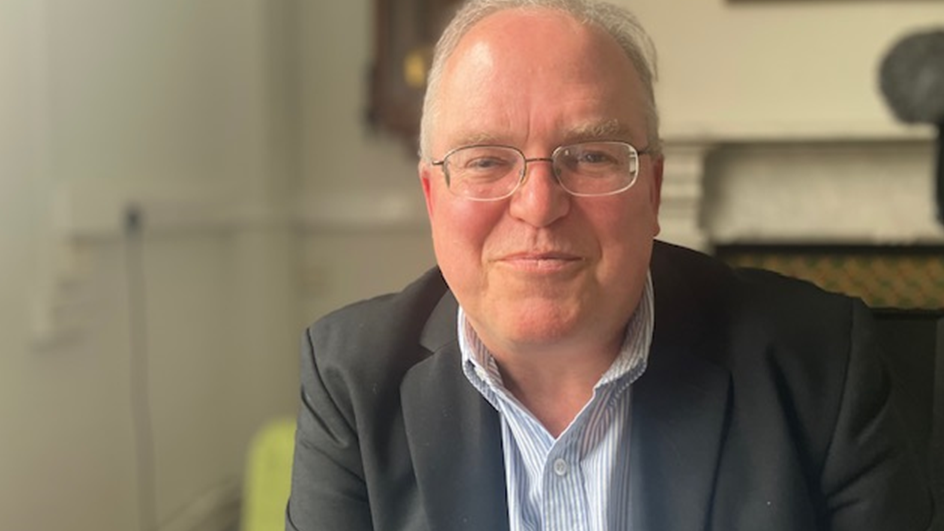 Dr Paul Cornes sitting wearing glasses and a suit looks at the camera