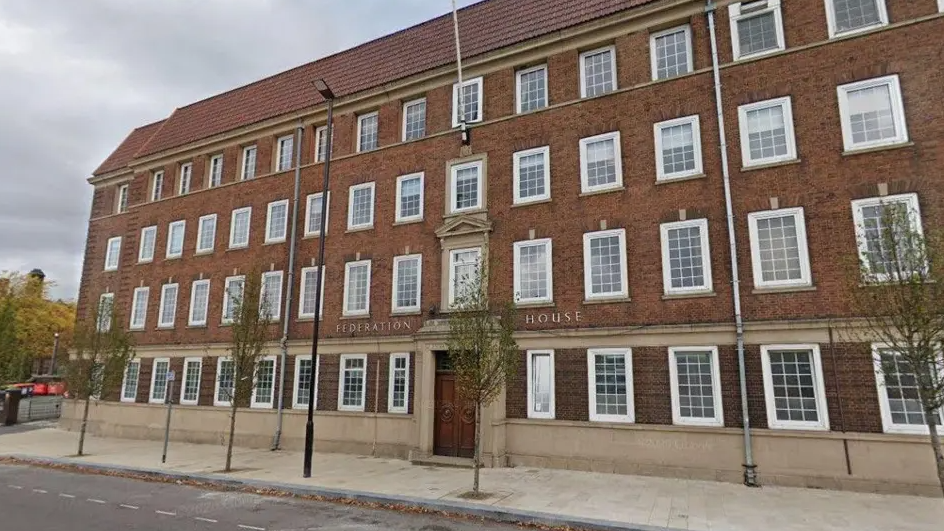 The large brick building with rows of windows has a front door in the centre and a pavement outside with a few small trees in front.