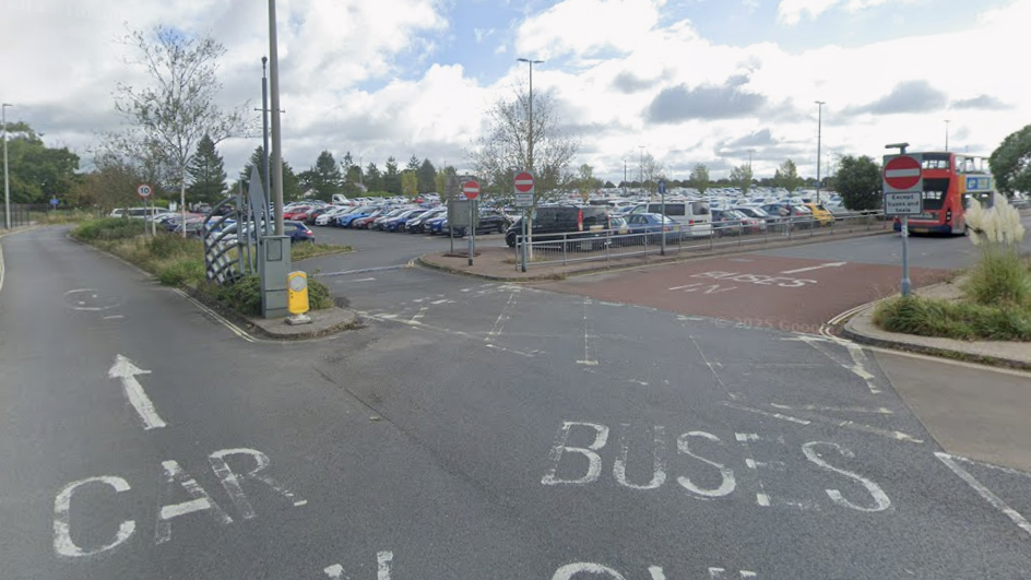 The entrance to the George Park and Ride. There are two lanes, the one on the left has an arrow leading straight ahead with the words CAP PARK painted on it while the other leads to the right and has BUSES with an arrow leading ahead painted on it. The car parking area are full of cars and there is a bus travelling parked on the side of the road.