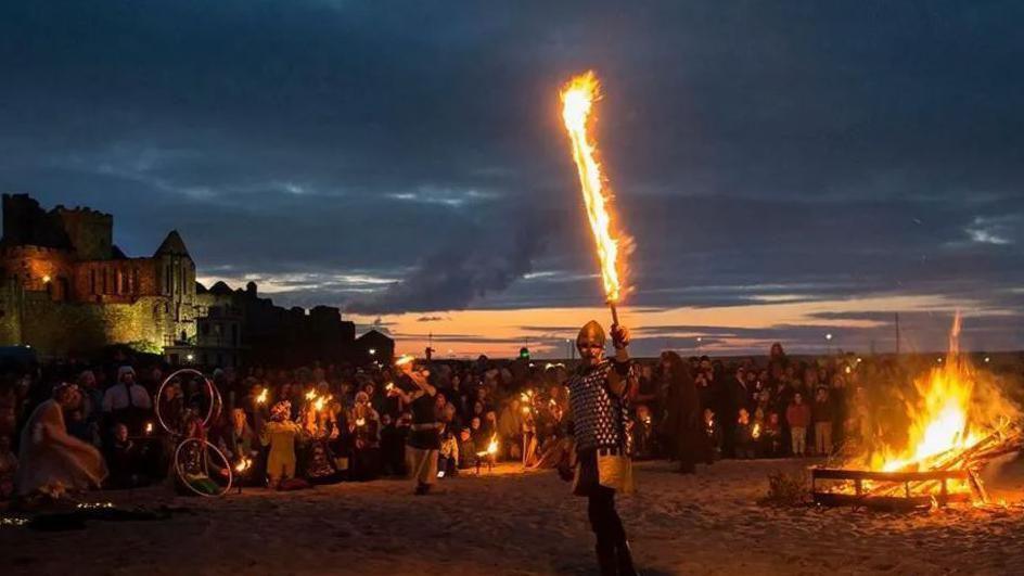 A man standing on Peel beach dressed as a Viking holding up a flaming sword in front of a crowd of people. There is a bonfire to the left and Peel Castle can bee seen in the background on the right.