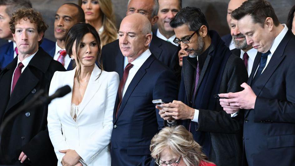 Inside the US Capitol Rotunda, a group of prominent tech and business leaders attend the inauguration ceremony of Donald Trump as the 47th President of the United States. From left to right: Meta CEO Mark Zuckerberg in a dark suit; Lauren Sánchez, wearing a white blazer over a lace bustier; Amazon founder Jeff Bezos in a navy suit; Google CEO Sundar Pichai in a dark suit and glasses; and Tesla and SpaceX CEO Elon Musk in a black suit and tie. They are standing or seated among other attendees in the ornate hall, with marble columns and historic paintings visible in the background.