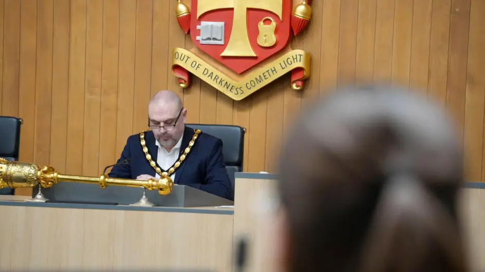 The council's deputy mayor Craig Collingswood is pictured in March sitting behind a desk at a council meeting with young people in front of him. He has his formal mayoral chain on over a navy suit. He has a grey beard and glasses on with a shaved head.


