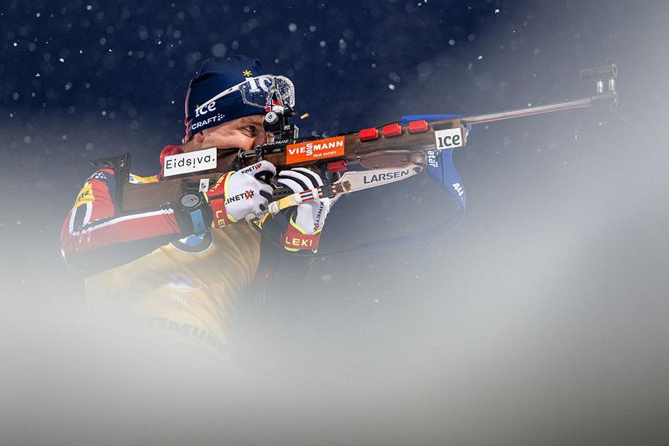 Johan-Olav Botn of Norway in winter gear aiming a rifle, with snow is blurred in the foreground, creating a dramatic image