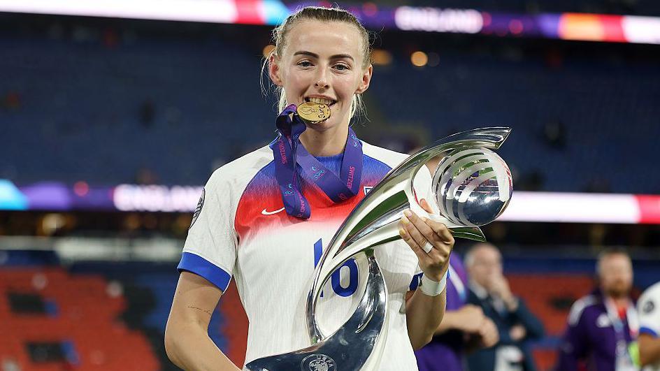 Chloe Kelly bites her winner’s medal while holding the UEFA Women’s Euro 2025 trophy, wearing her England kit on the pitch after the final.
