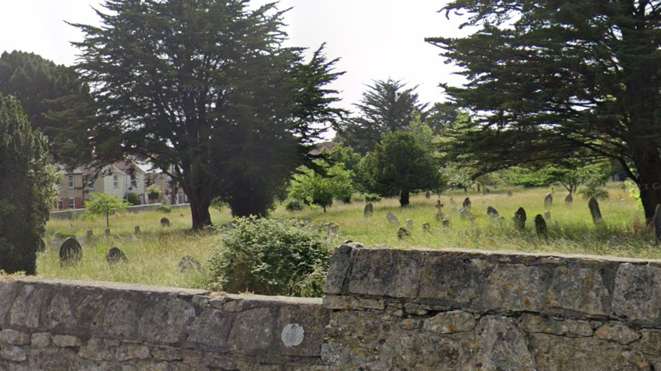 A cemetery with dozens of gravestones. Grass surrounding them has not been cut and is overgrown