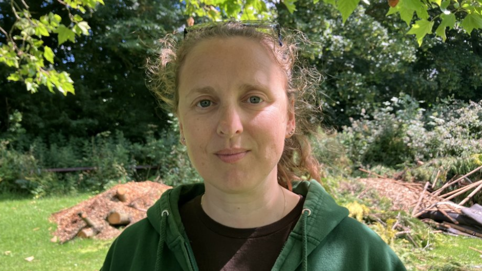 Jemmima Woods is wearing a green hoodie and standing in front of a large pile of logs and wood chippings