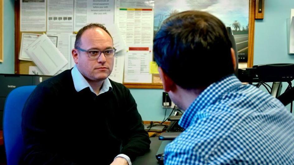 Dr Jamie Green faces a male patient in his surgery. He is wearing a light-coloured open-necked shirt over a dark sweater. He has short hair and glasses. Behind him is a pinboard and the back of a computer monitor. The patient is wearing a gingham shirt and has dark hair. He is facing away from the camera.