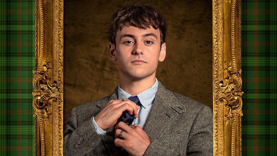 The image shows Tom Daley in a formal suit and tie, adjusting his tie. He is framed by an ornate golden frame with green tartan-patterned sides. The background features a textured brown surface.