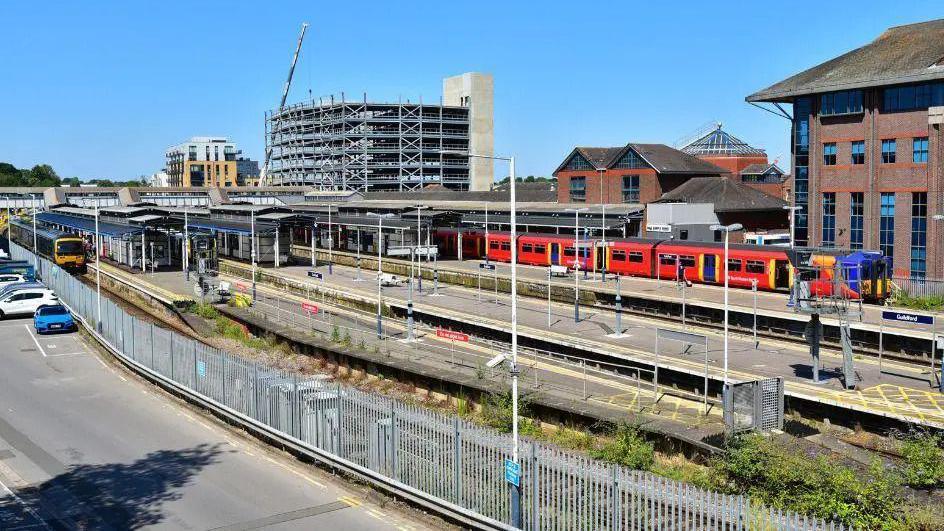 Guildford Railway Station. There is clear blue skies, a red train stopped at the station and a car park.