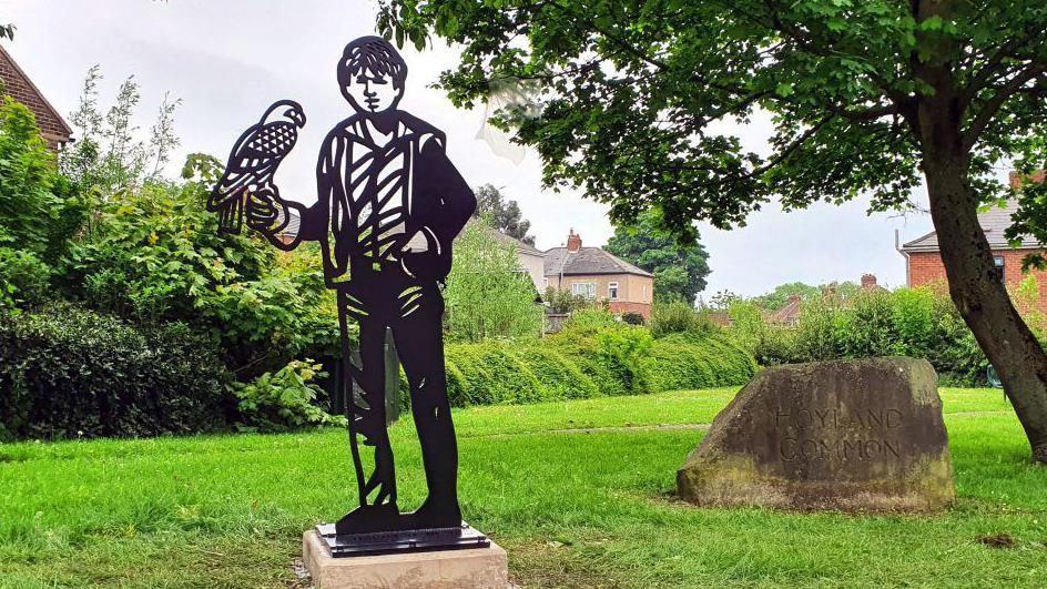 A steel sculpture of a boy holding a kestrel set in an open area of Hoyland Common, based on the book 'A Kestrel for a Knave' by Barry Hines. 