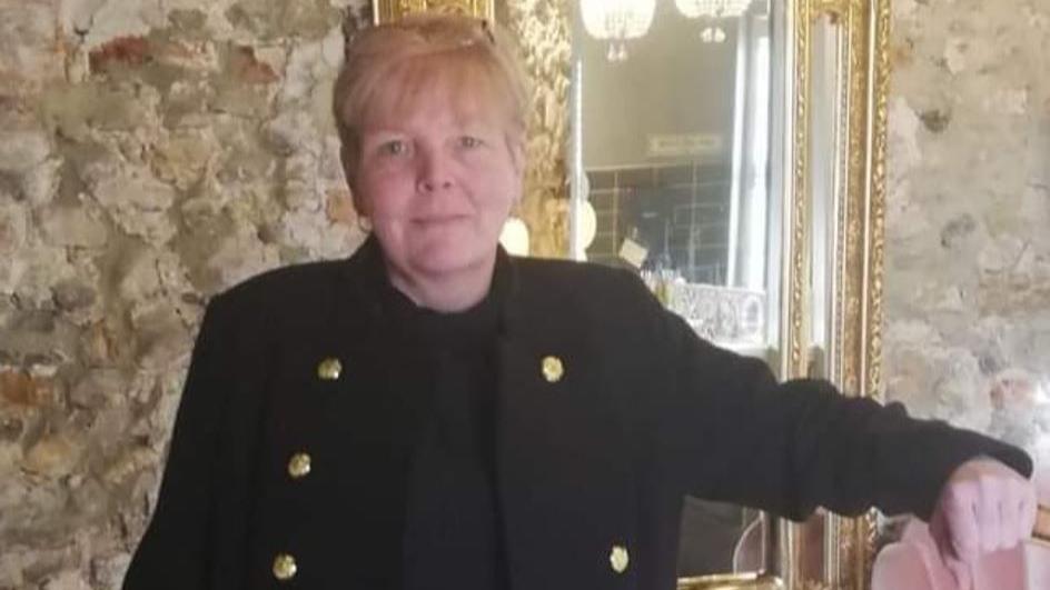 Picture of Eve McIntyre. She has short fair hair in a fringe, wearing a black jumper and large black coat with large gold buttons. She is standing in front of a stone wall and a mirror with a gold frame.