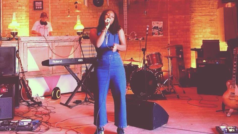 A woman is wearing a denim jumpsuit and is standing in a studio filled with musical instruments and orange mood lighting. 