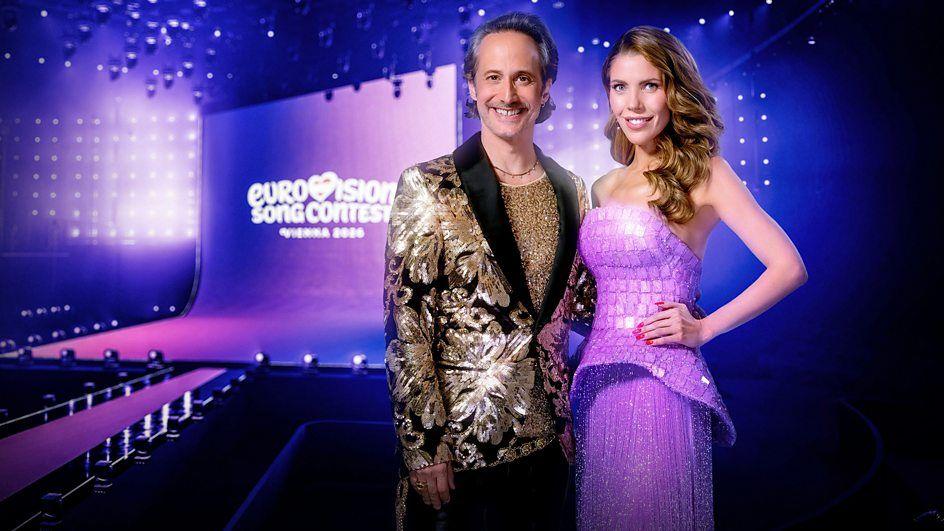 Victoria Swarovski and Michael Ostrowski Stand in front of a screen showing the Eurovision logo