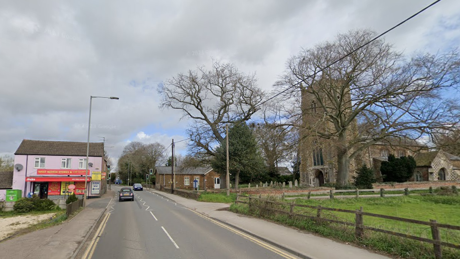 Google map view of the road through West Winch. There is a road and on the left side a pink house and the right hand side a church and grassed field. 