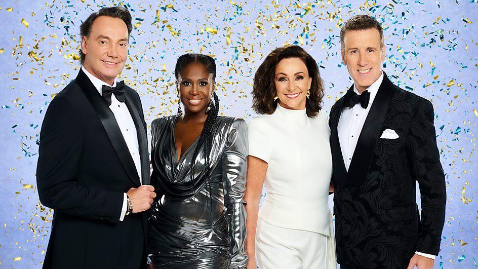 From left to right Craig Revel Horwood, Motsi Mabuse, Head Judge Shirley Ballas and Anton Du Beke can be seen standing in front of a blue background with glitter in the air around them. 