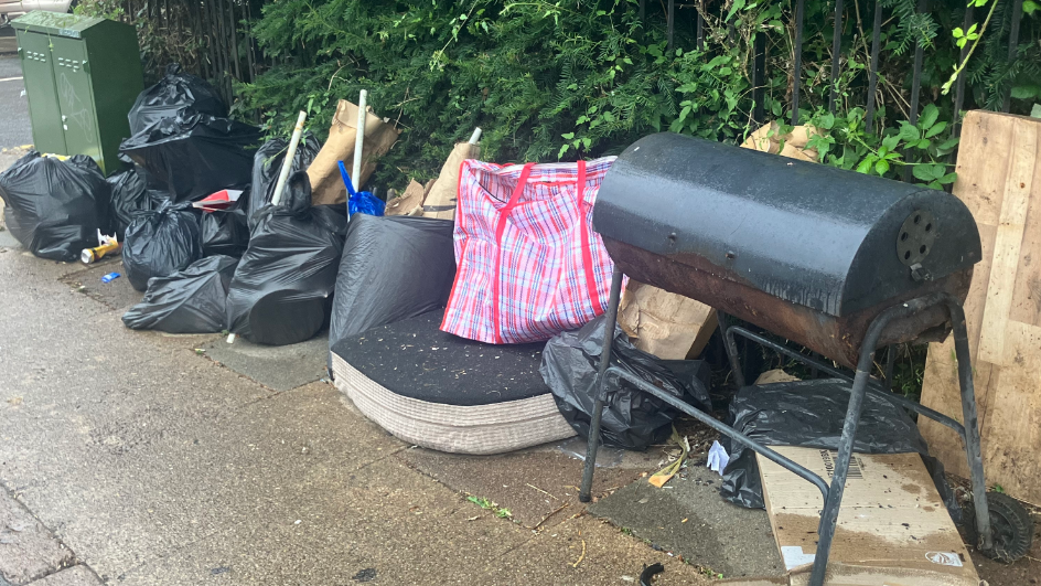 Rubbish dumped on Semilong Road, Northampton. It includes a black barbeque on legs, black plastic bin bags with cardboard sticking out, and a seat cushion.